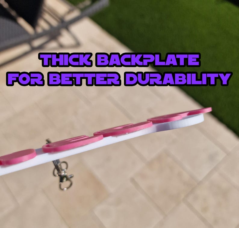 May include: A white and pink keychain with the text "THICK BACKPLATE FOR BETTER DURABILITY". The keychain has several pink circular accents and a silver clasp. The design is set against a blurred outdoor background.