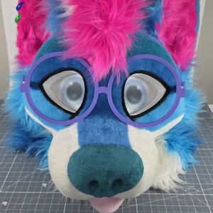 May include: A colorful furry head with blue, pink, and white fur. The head has large, expressive eyes and is wearing purple-rimmed glasses. The nose is teal, and the tongue is pink. The head is designed to resemble a cartoon animal.