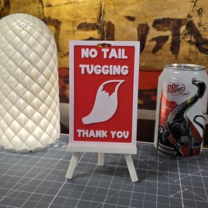 K&ouml;nnte beinhalten: Ein rotes Schild mit wei&szlig;em Text, der "NO TAIL TUGGING" und "THANK YOU" lautet. Das Schild zeigt eine wei&szlig;e Fuchsschwanz-Grafik und wird auf einer kleinen wei&szlig;en Staffelei angezeigt. Eine Dr. Pepper-Dose ist im Hintergrund zu sehen.