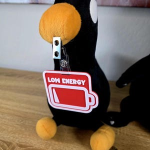 May include: A black plush penguin with an orange beak and feet. It has a white eye and a red badge that says "LOW ENERGY" with a battery icon. The badge is attached with a metal clip. The penguin is sitting.