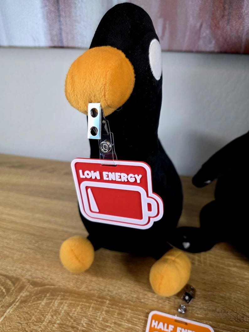 May include: A black plush penguin with an orange beak and feet. It has a white eye and a red badge that says "LOW ENERGY" with a battery icon. The badge is attached with a metal clip. The penguin is sitting.