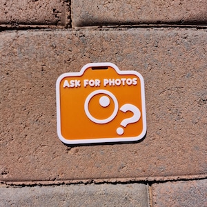 May include: An orange camera-shaped sticker with white trim and the text "ASK FOR PHOTOS". The design includes a white circle and a question mark. The sticker is placed on a textured, reddish-brown brick surface.