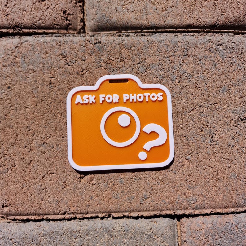 May include: An orange camera-shaped sticker with white trim and the text "ASK FOR PHOTOS". The design includes a white circle and a question mark. The sticker is placed on a textured, reddish-brown brick surface.