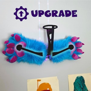 May include: A pair of blue and pink furry paw-shaped accessories with the word "UPGRADE" above them. The paws have pink pads and claws, and are connected by a black structure. The background is a white wall.