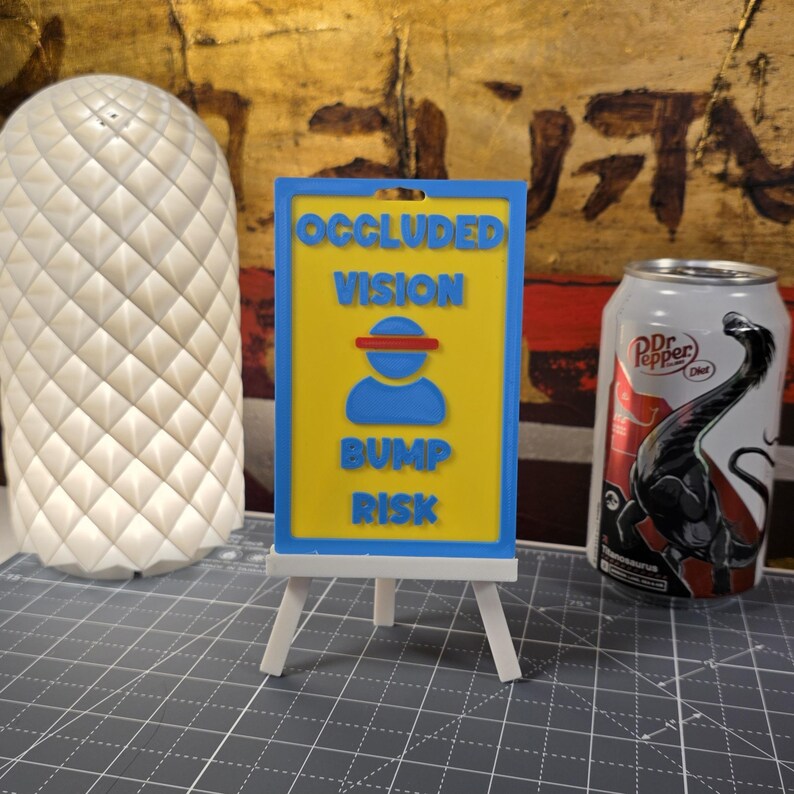 K&ouml;nnte beinhalten: Ein 3D-gedrucktes Schild auf einer kleinen Staffelei mit den Worten "OCCLUDED VISION BUMP RISK" in Blau und Gelb. Eine wei&szlig;e, strukturierte Lampe und eine Dr. Pepper-Dose mit einer Dinosaurier-Grafik sind ebenfalls im Bild.