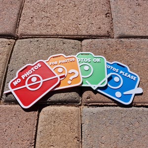 May include: Four colorful camera-shaped signs with white outlines. The signs are red, orange, green, and blue, each with a different message: "NO PHOTOS", "FOR PHOTOS", "PHOTOS OK", and "PHOTOS PLEASE".