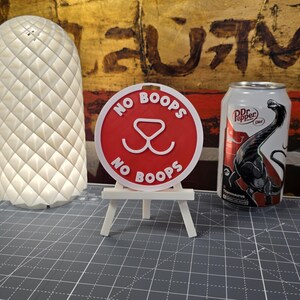 K&ouml;nnte beinhalten: Ein rotes rundes Schild mit den Worten "NO BOOPS" und einer Hunde-Nasen-Grafik, das auf einer kleinen wei&szlig;en Staffelei ausgestellt ist. Rechts ist eine Dose Dr. Pepper Diet. Links ist eine wei&szlig;e, strukturierte Lampe.