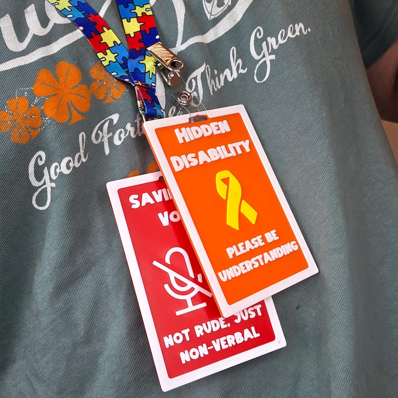 Op de afbeelding: Een lanyard met een kleurrijk puzzelstukjespatroon en een metalen clip met twee rechthoekige badges. E&eacute;n badge is oranje met de woorden "HIDDEN DISABILITY" en een gele lint. De andere is rood met een microfoonsymbool en de woorden "NOT RUDE, JUST NON-VERBAL."