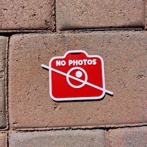 May include: A red and white sign with the words "NO PHOTOS" and a crossed-out camera icon. The sign is rectangular with rounded corners and is placed on a brick surface.