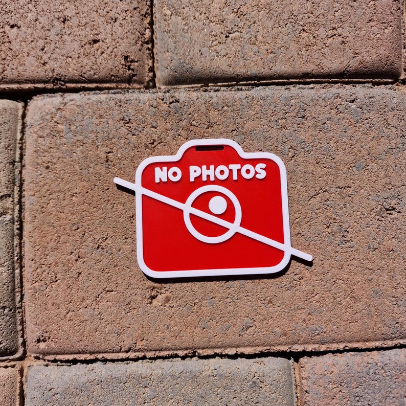 May include: A red and white sign with the words "NO PHOTOS" and a crossed-out camera icon. The sign is rectangular with rounded corners and is placed on a brick surface.