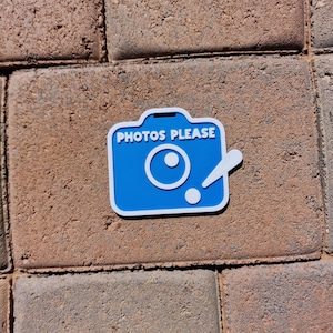 May include: A blue and white camera-shaped sticker with the text "PHOTOS PLEASE". The sticker is on a brick surface. The camera design includes a lens and a flash.