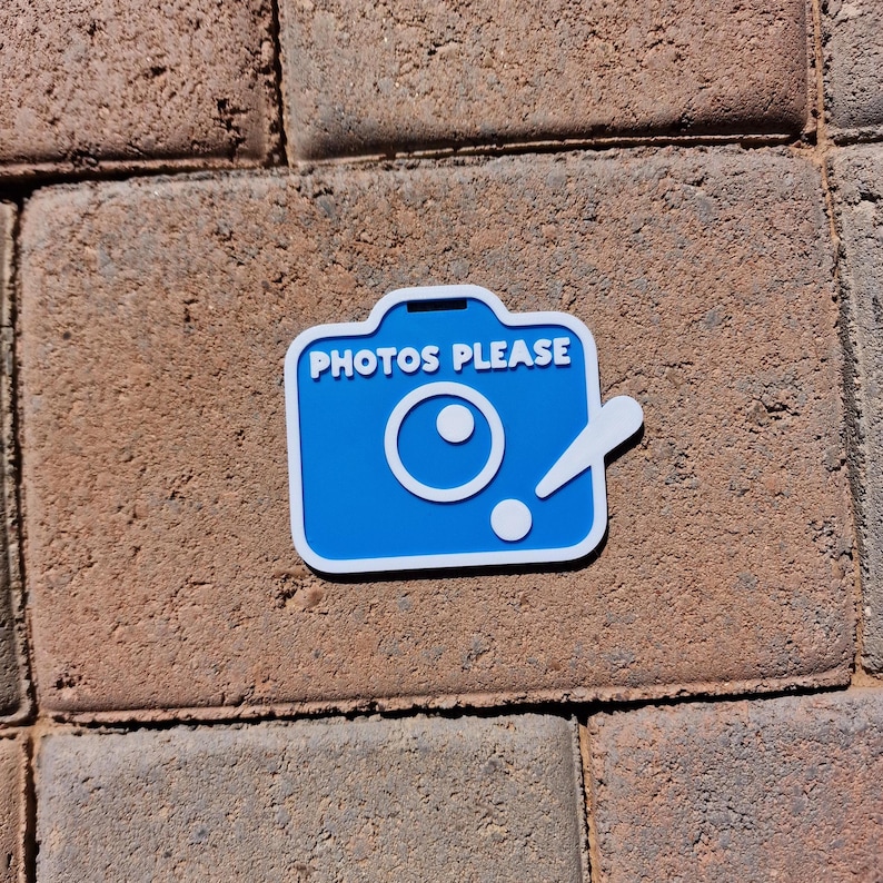 May include: A blue and white camera-shaped sticker with the text "PHOTOS PLEASE". The sticker is on a brick surface. The camera design includes a lens and a flash.