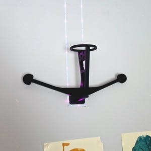 May include: A black, wall-mounted decorative piece with a circular top and two arms extending outwards, each ending in a circular shape. The piece has a rectangular base and a vertical support with text. The wall is white.