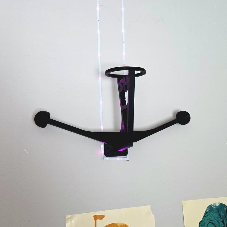 May include: A black, wall-mounted decorative piece with a circular top and two arms extending outwards, each ending in a circular shape. The piece has a rectangular base and a vertical support with text. The wall is white.
