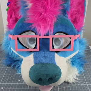 May include: A furry blue and white wolf head with pink accents, including a pink mohawk and pink square glasses. The wolf has large, expressive eyes and a teal nose. The mouth is slightly open, revealing a pink tongue.