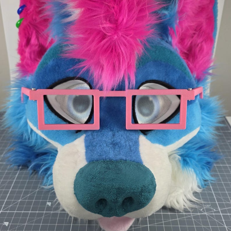 May include: A furry blue and white wolf head with pink accents, including a pink mohawk and pink square glasses. The wolf has large, expressive eyes and a teal nose. The mouth is slightly open, revealing a pink tongue.