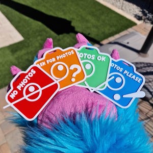 May include: Four colorful camera-shaped signs with text: "NO PHOTOS", "ASK FOR PHOTOS", "PHOTOS OK", and "PHOTOS PLEASE". The signs are red, orange, green, and blue, with white text and camera lens designs. The signs are held by a furry, blue and pink creature.