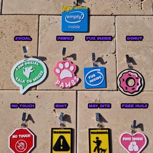 May include: A collection of colorful, novelty tags with various designs and text. Includes a blue tag with "(empty) inside", a pink paw print tag with "PAWBS", and a red "NO TOUCH" tag. Other tags have phrases like "MAY BITE" and "FREE HUGS!"