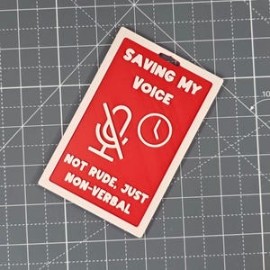 Puede incluir: Una insignia rectangular roja y blanca con el texto "SAVING MY VOICE" y "NOT RUDE, JUST NON-VERBAL". La insignia presenta un micr&oacute;fono tachado y un icono de reloj.