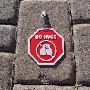 Op de afbeelding: Een rood en wit achthoekig bord met de woorden "NO HUGS" in witte tekst. Het bord heeft een witte afbeelding van twee figuren die elkaar omarmen, doorgestreept met een rode lijn. Een metalen clip is aan de bovenkant bevestigd.