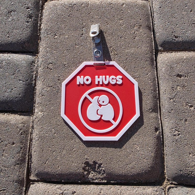 Op de afbeelding: Een rood en wit achthoekig bord met de woorden "NO HUGS" in witte tekst. Het bord heeft een witte afbeelding van twee figuren die elkaar omarmen, doorgestreept met een rode lijn. Een metalen clip is aan de bovenkant bevestigd.