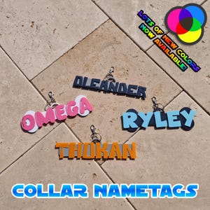 May include: A collection of colorful collar nametags with names like "Omega," "Oleander," and "Ryley." The tags are in various colors, including pink, blue, and orange, with a keychain attachment. Text in the image reads "Lots of New Colors Now Available!"