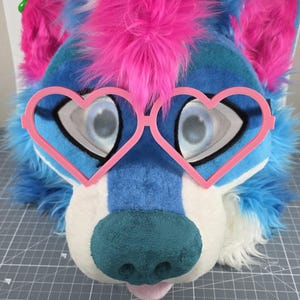 May include: A furry wolf head with blue and white fur, pink accents, and heart-shaped pink glasses. The wolf has a teal nose and a pink tongue. The eyes are large and detailed.
