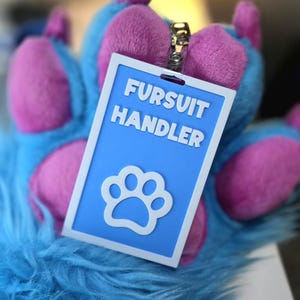May include: A blue and white rectangular badge with the text "FURSUIT HANDLER" and a paw print symbol. The badge is attached to a silver clip and is set against a furry blue and purple background.