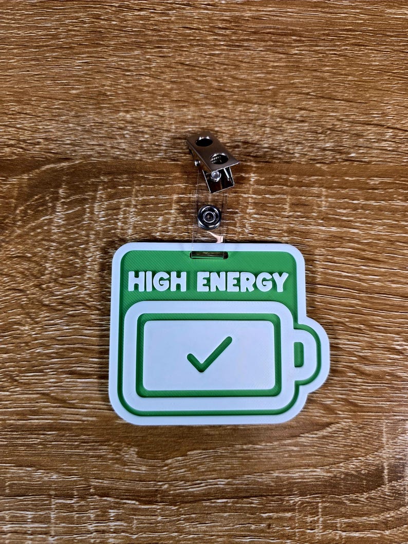 May include: A green and white badge with the words "HIGH ENERGY" and a battery icon with a checkmark. The badge has a metal clip for attachment.