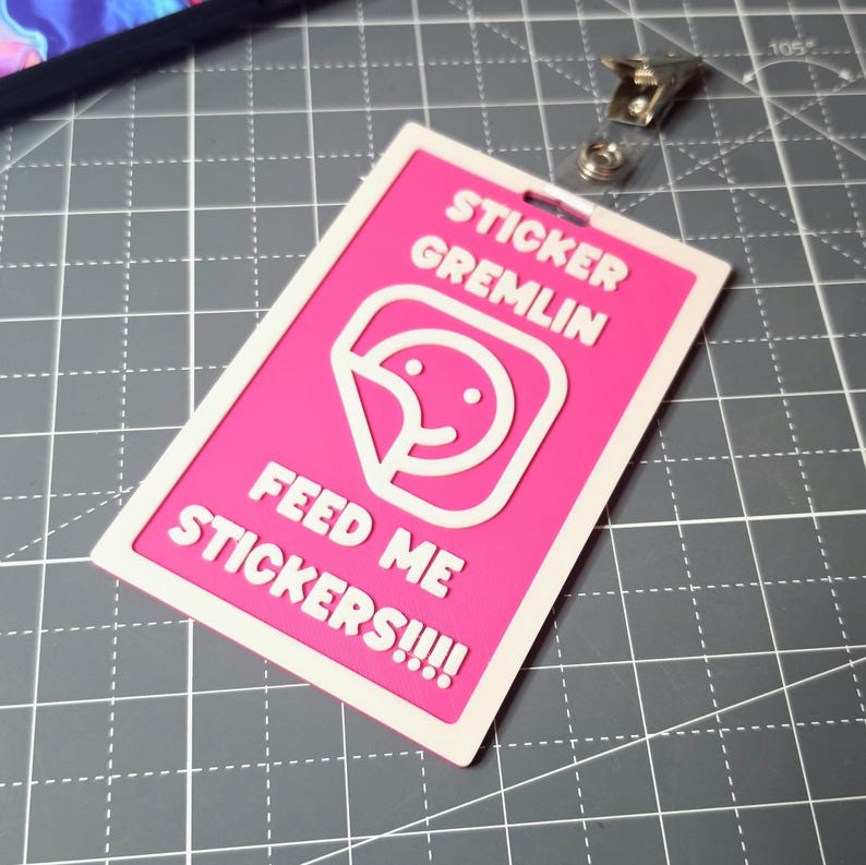 May include: A pink and white rectangular sticker with the text "STICKER GREMLIN" and "FEED ME STICKERS!!!!" in white. The sticker features a white graphic of a smiling face with a peeled sticker. A metal clip is attached.
