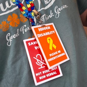May include: A lanyard with a colorful puzzle piece pattern and a metal clip holds two rectangular badges. One badge is orange with the words "HIDDEN DISABILITY" and a yellow ribbon. The other is red with a microphone symbol and the words "NOT RUDE, JUST NON-VERBAL."