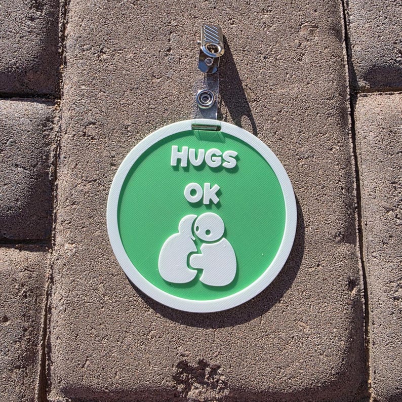 Op de afbeelding: Een ronde, groen-witte badge met de witte tekst "HUGS OK". Onder de tekst staat een witte afbeelding van twee figuren die elkaar omhelzen. De badge is bevestigd aan een metalen clip.