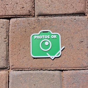 May include: A green camera-shaped sticker with the text "PHOTOS OK" in white. The sticker has a white border and a white lens design. The sticker is placed on a brick surface.