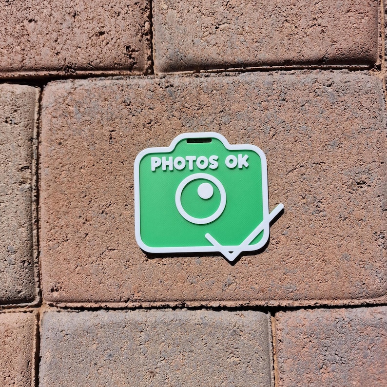 May include: A green camera-shaped sticker with the text "PHOTOS OK" in white. The sticker has a white border and a white lens design. The sticker is placed on a brick surface.