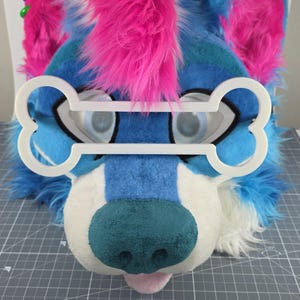 May include: A plush, furry dog head in shades of blue, white, and teal, with pink accents. The dog has a teal nose and a pink tongue. A white bone-shaped frame sits over the eyes. The head is likely part of a costume or cosplay.