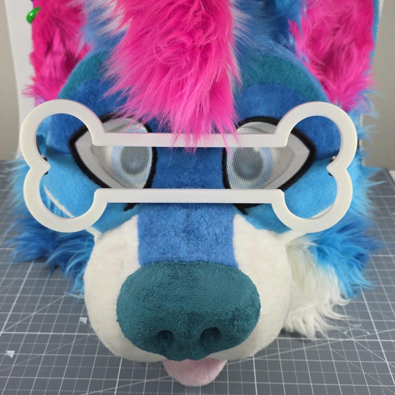 May include: A plush, furry dog head in shades of blue, white, and teal, with pink accents. The dog has a teal nose and a pink tongue. A white bone-shaped frame sits over the eyes. The head is likely part of a costume or cosplay.