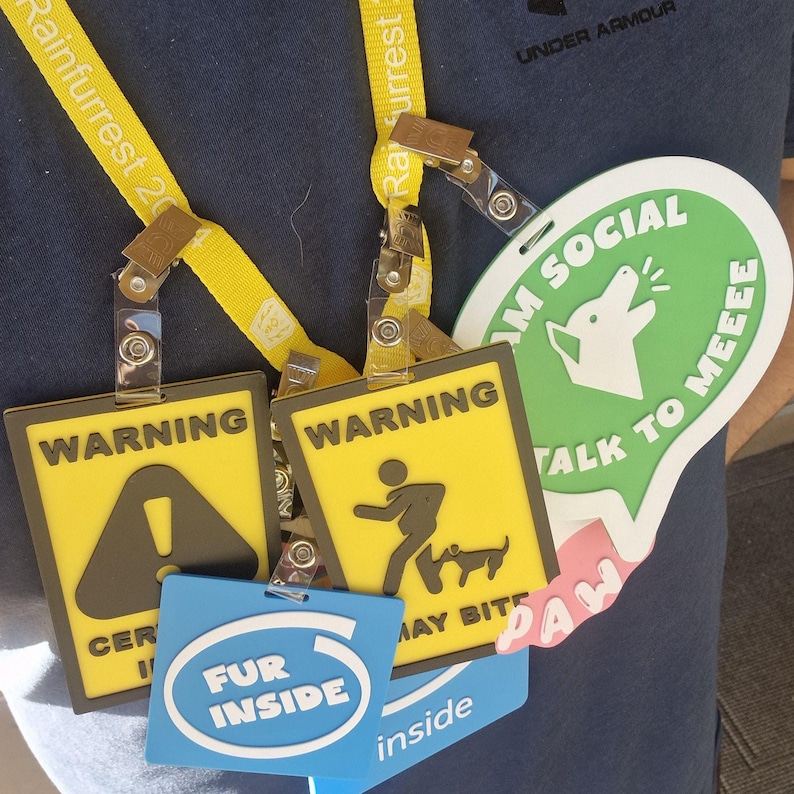 May include: A collection of event badges and lanyards. Two yellow and black warning signs with the text "WARNING" and a caution symbol and a person walking a dog. A green badge says "I'M SOCIAL" and "TALK TO MEEEE". A blue badge says "FUR INSIDE".