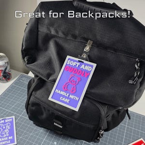 Puede incluir: Una mochila negra con una etiqueta que dice "SOFT AND CUDDLY" con un gr&aacute;fico de oso de peluche rosa. La etiqueta tambi&eacute;n dice "HANDLE WITH CARE". El texto "Great for Backpacks!" est&aacute; en la parte superior.