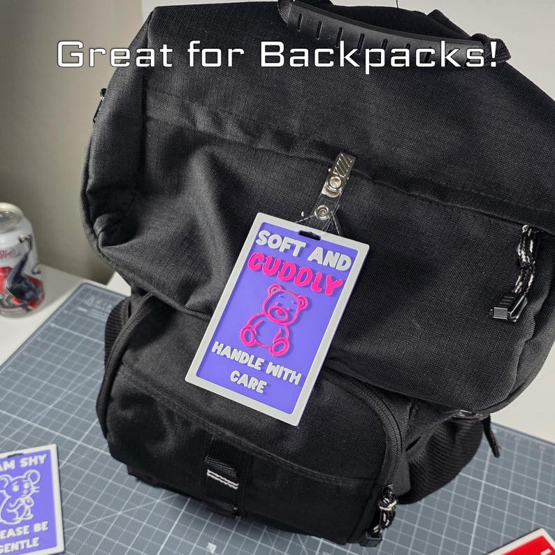 Puede incluir: Una mochila negra con una etiqueta que dice "SOFT AND CUDDLY" con un gr&aacute;fico de oso de peluche rosa. La etiqueta tambi&eacute;n dice "HANDLE WITH CARE". El texto "Great for Backpacks!" est&aacute; en la parte superior.