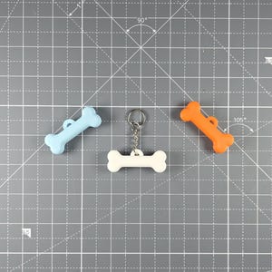 May include: Three dog bone-shaped keychains in light blue, white, and orange. The white bone is attached to a silver keyring. Each bone has a small hole for attachment. The keychains are on a grey cutting mat.