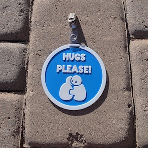 Op de afbeelding: Een ronde, blauw-witte bord met de witte tekst "HUGS PLEASE!". Onder de tekst staat een afbeelding van twee figuren die elkaar omhelzen. Het bord is bevestigd aan een doorzichtige plastic clip.