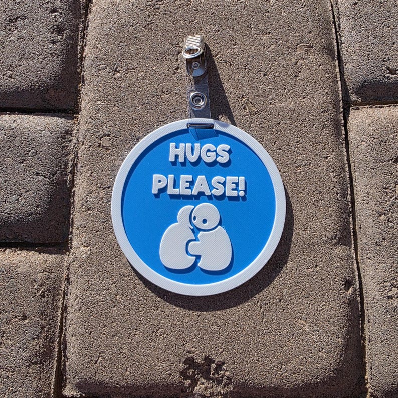 Op de afbeelding: Een ronde, blauw-witte bord met de witte tekst "HUGS PLEASE!". Onder de tekst staat een afbeelding van twee figuren die elkaar omhelzen. Het bord is bevestigd aan een doorzichtige plastic clip.