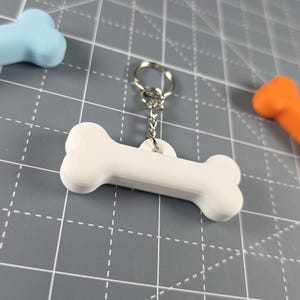 May include: A white, bone-shaped keyring with a silver ring. The keyring is the main focus, with two other bone-shaped objects in blue and orange in the background. The objects are on a grey surface.