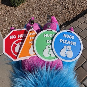 Op de afbeelding: Een kleurrijke pluche poot met borden die de knuffelvoorkeuren aangeven. De borden, in rood, oranje, groen en blauw, luiden "Geen knuffels", "Vraag om knuffels", "Knuffels OK" en "Knuffels graag!".