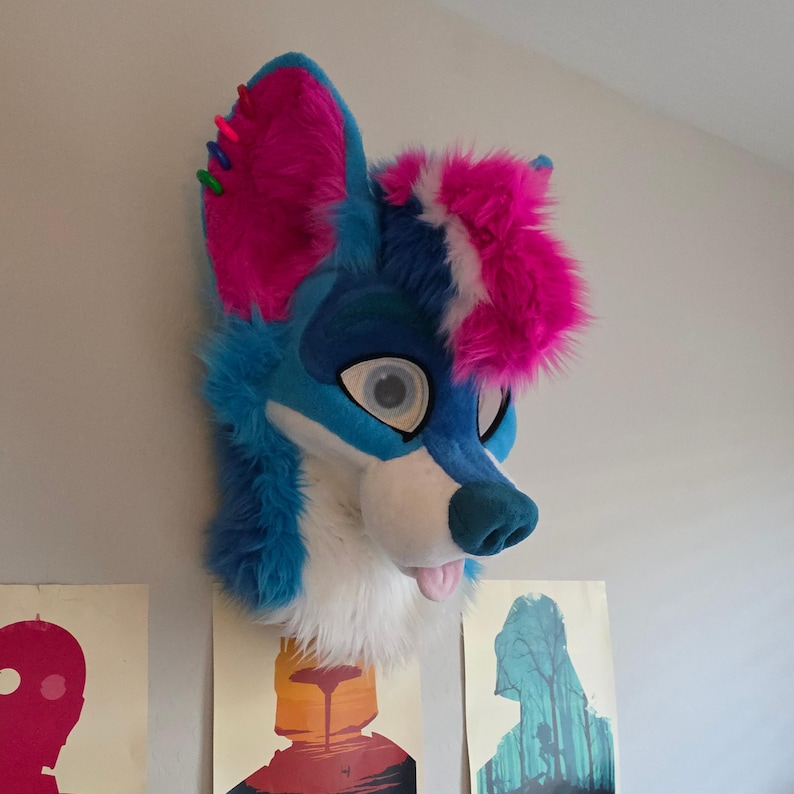 May include: A blue, white, and pink furry fox head with large, round eyes and a protruding tongue. The fox head has a blue nose and ears with pink accents. The head is mounted on a wall, with colorful posters visible below.