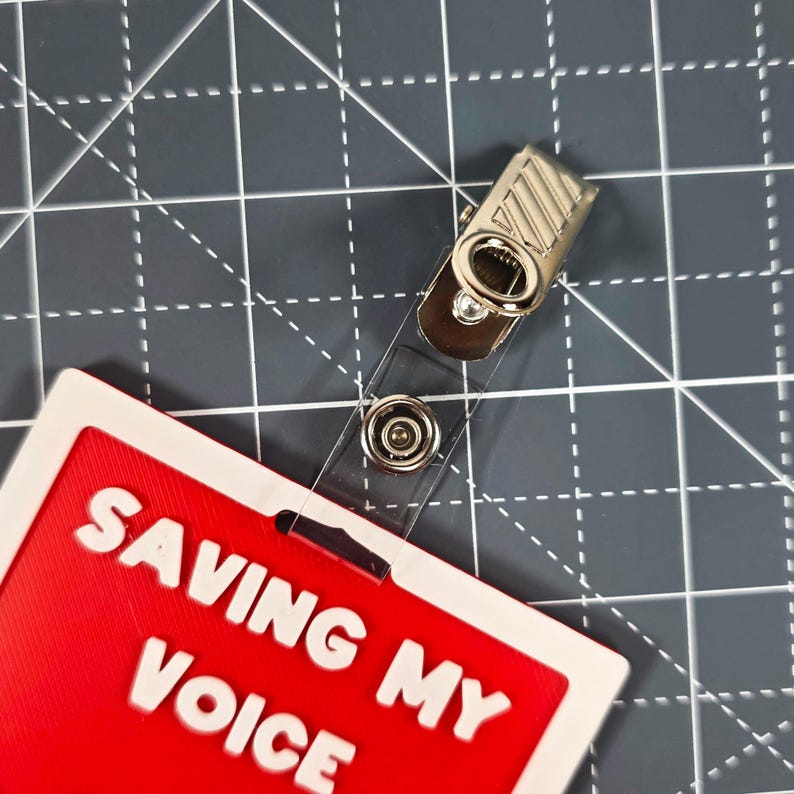 Puede incluir: Una placa de identificaci&oacute;n roja y blanca con el texto "SAVING MY VOICE" en blanco. La placa est&aacute; sujeta a una correa de pl&aacute;stico transparente con un clip met&aacute;lico.