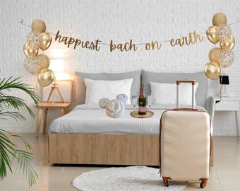 May include: A bedroom scene with a bed, balloons, and a suitcase. Gold balloons and a banner that reads "happiest bach on earth" decorate the room. A champagne bottle and glasses sit on a tray. A beige suitcase with brown accents is in the foreground.