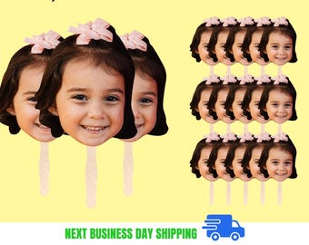 May include: A collection of face cutouts of a smiling child with brown hair and a pink bow. The cutouts are attached to wooden sticks. The background is a solid yellow color.