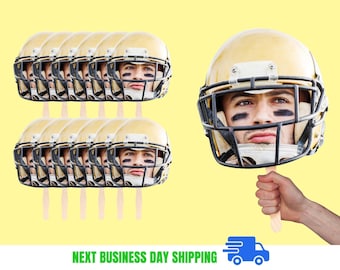 May include: A collection of football helmet face cutouts on wooden sticks. The helmets are gold with black face masks, and the faces have black eye paint. The background is a light yellow color.