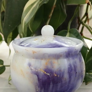 May include: A small, round, lidded jar with a white and purple marbled design. The jar has a small, round white knob on top of the lid. Gold flecks are scattered across the surface. The jar is set against a backdrop of green foliage.